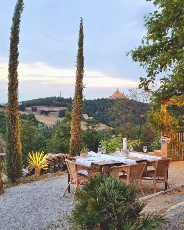 Tavolo apparecchiato tra i cipressi al tramonto, con il Santuario di San Luca sui colli bolognesi